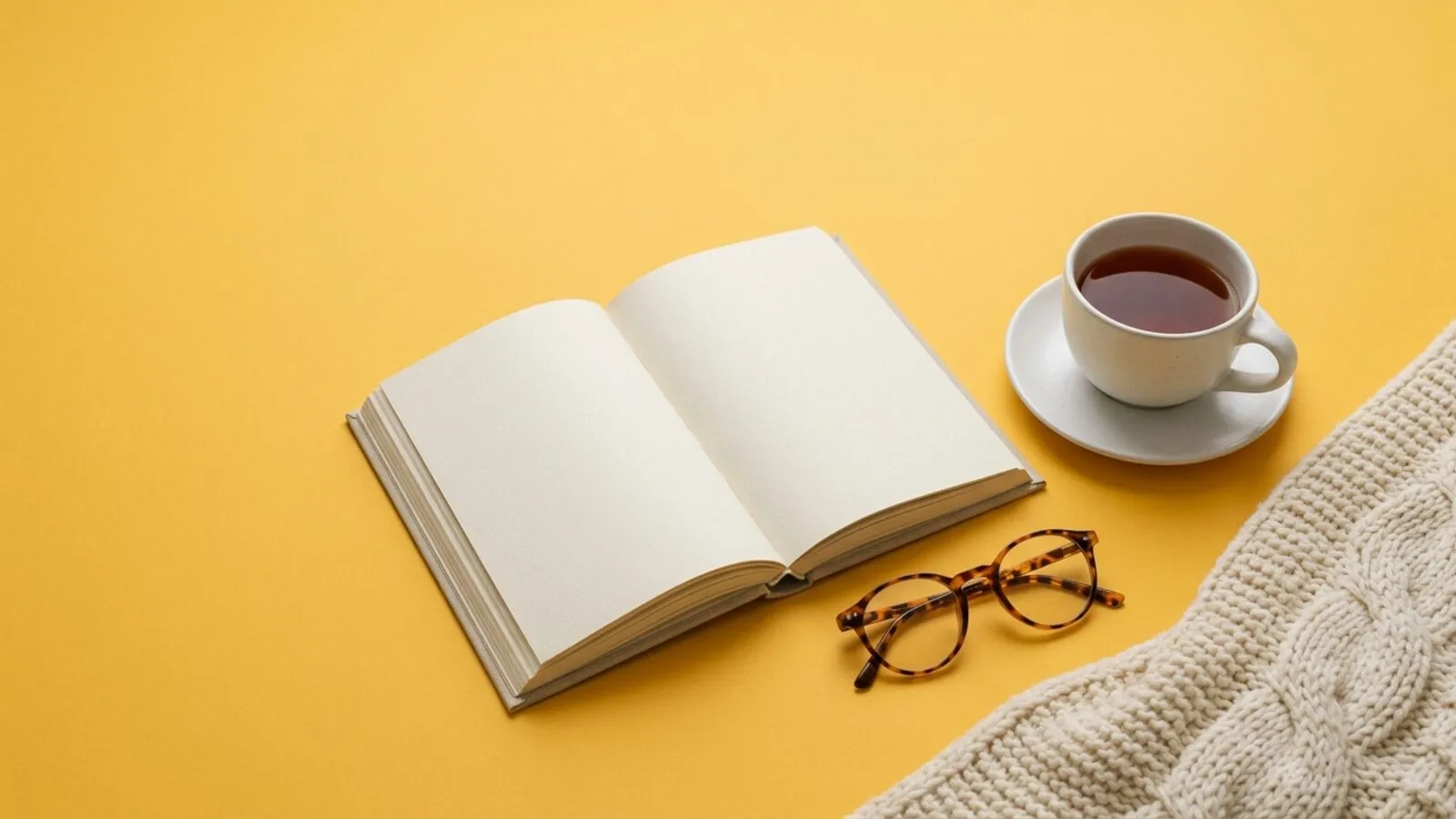 Livre ouvert aux pages crème, lunettes écaille, tasse de thé et coin de plaid en maille torsadée sur fond jaune