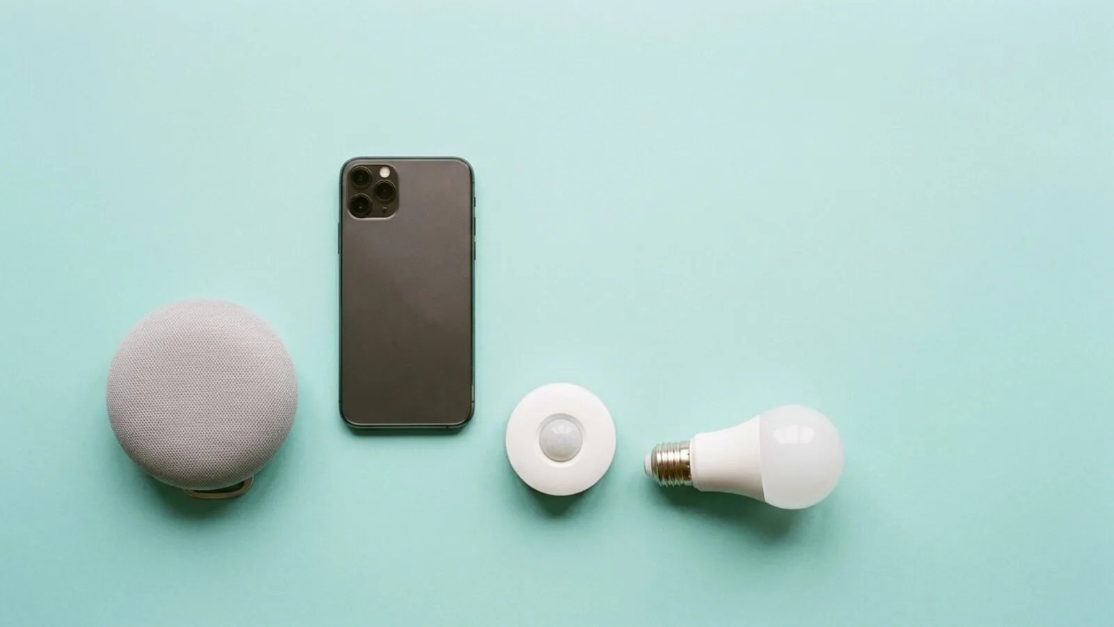 Enceinte connectée en tissu gris, smartphone, détecteur de mouvement rond et ampoule blanche sur fond bleu-vert
