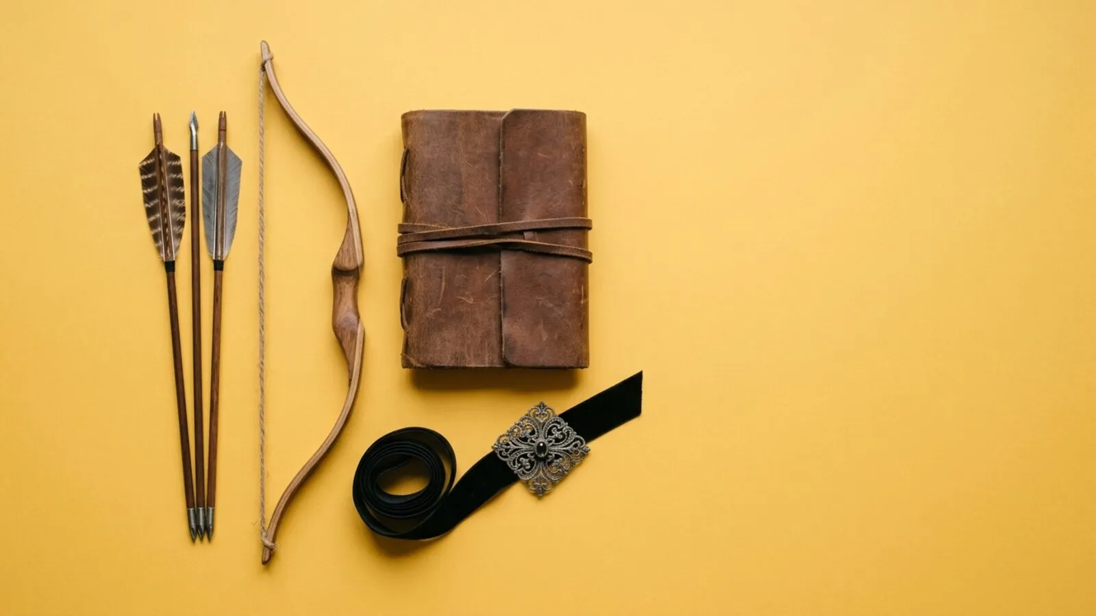 Arc en bois et trois flèches, carnet en cuir, ruban de velours noir et broche ancienne en argent sur fond jaune