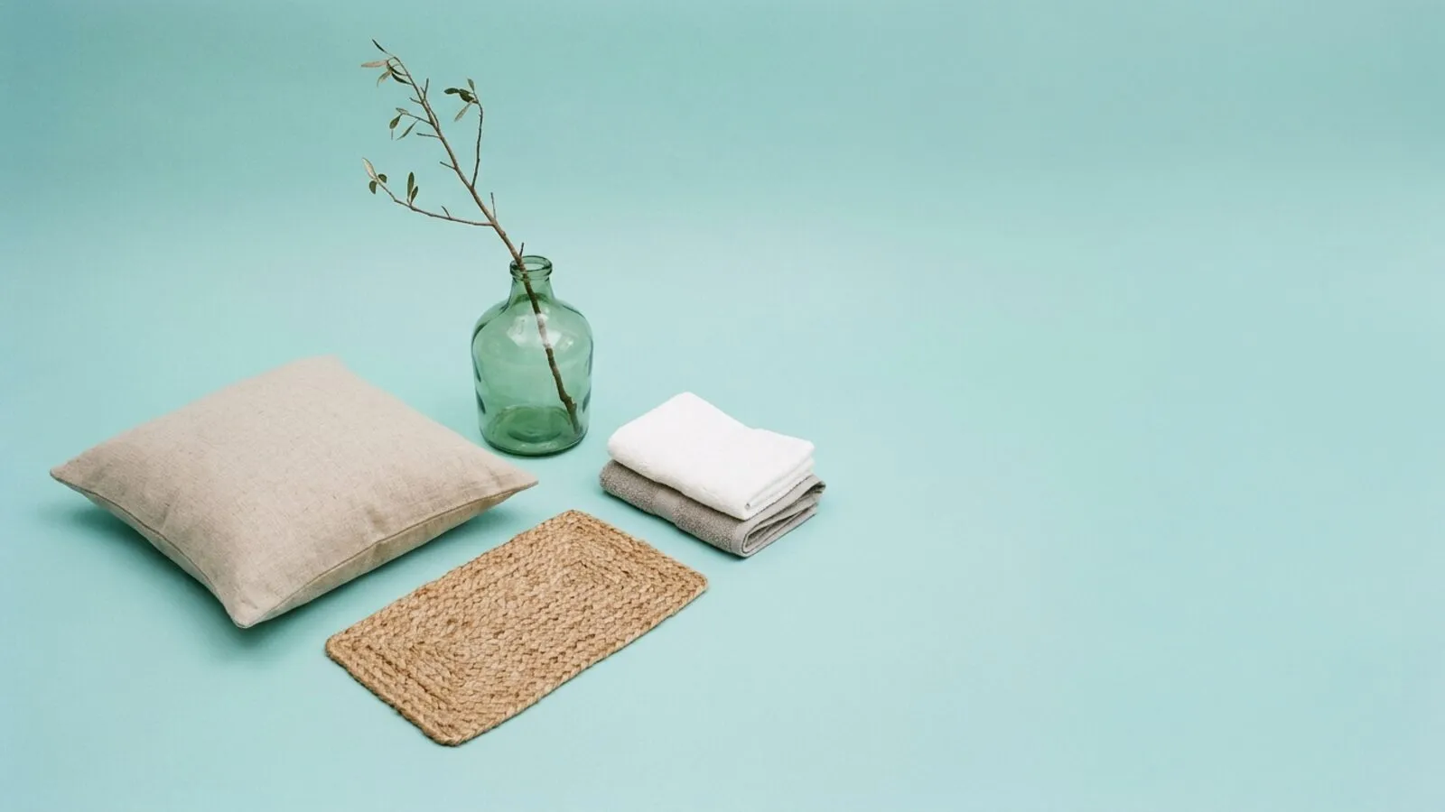 Housse de coussin en lin, tapis en jute, vase en verre recyclé avec une branche et serviettes pliées sur fond bleu-vert