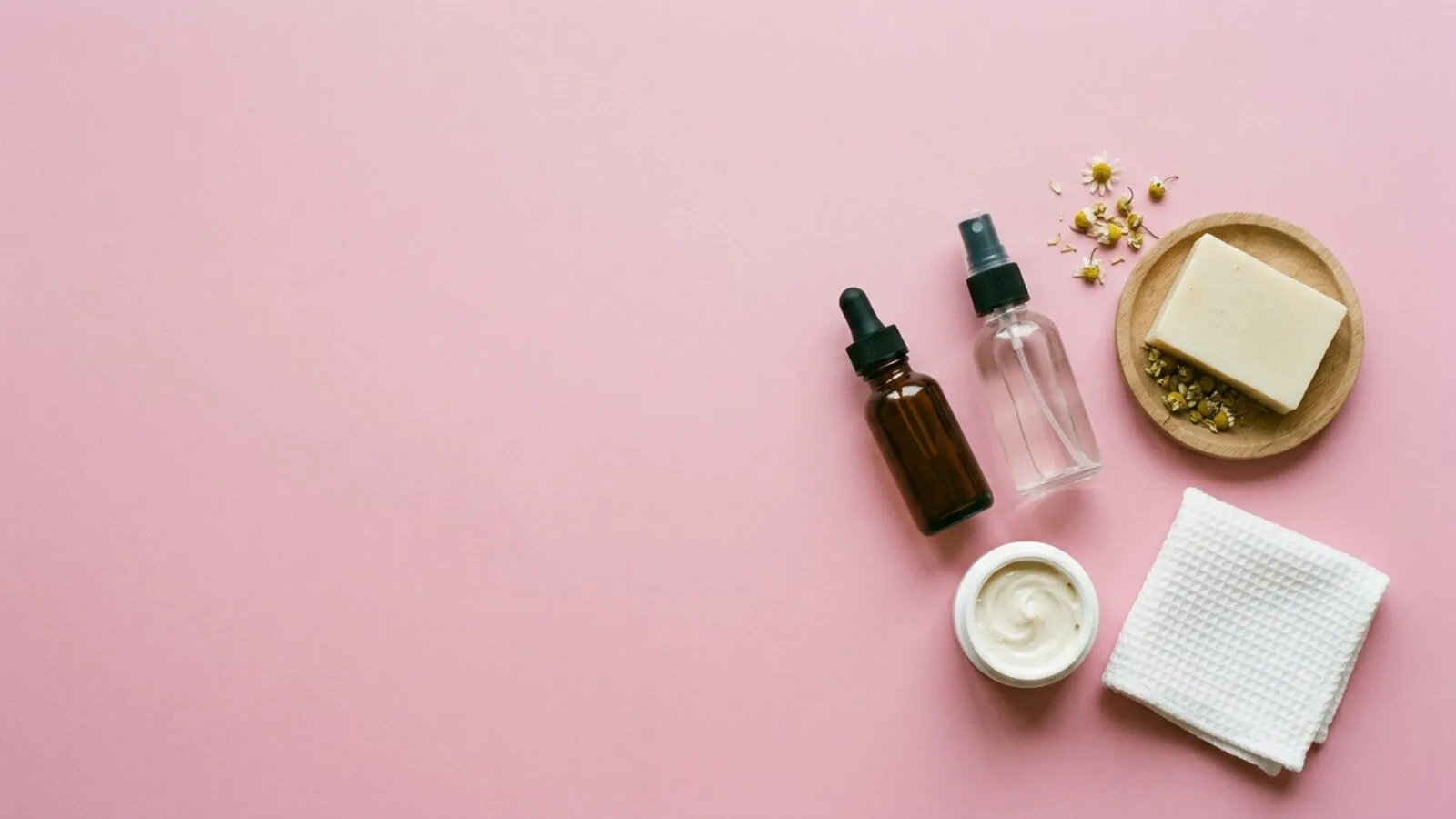 Flacon compte-gouttes ambré, vaporisateur, pot de crème, savon sur coupelle en bois et camomille sur fond rose