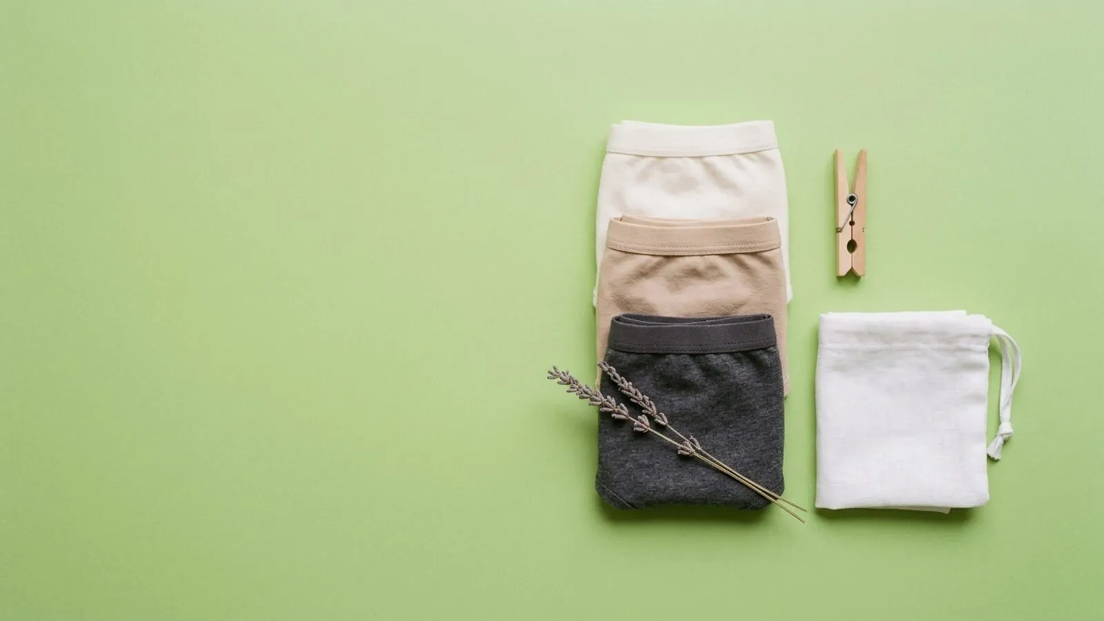 Trois culottes en coton écru, beige et gris pliées, pince à linge, pochette en lin et lavande sur fond vert tendre
