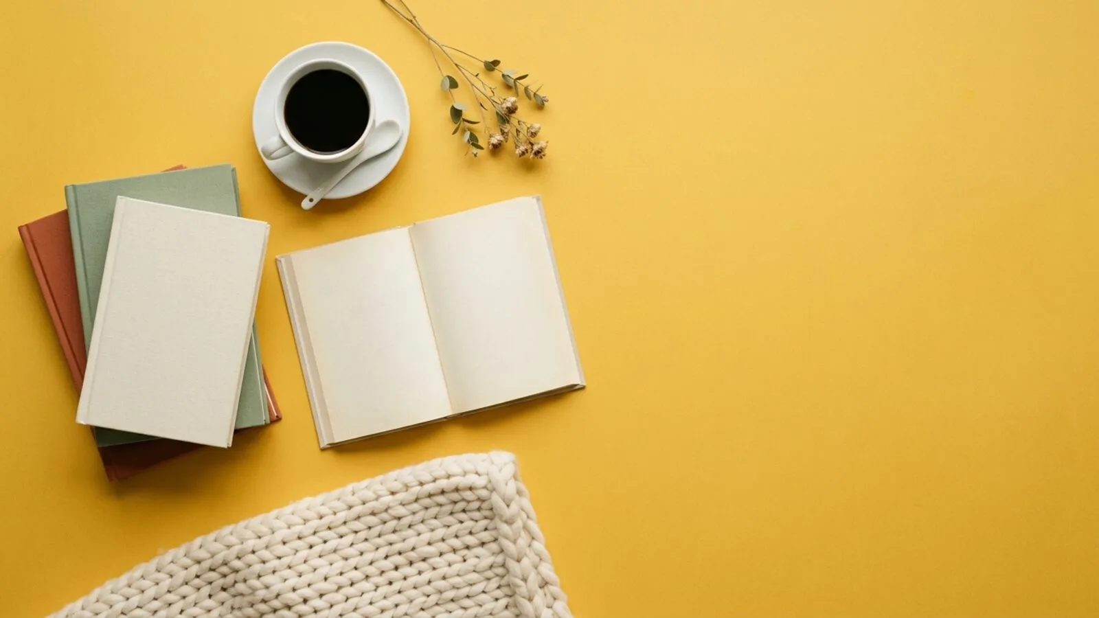 Livres aux couvertures unies, livre ouvert, tasse de café noir, brin de fleurs séchées et plaid crème sur fond jaune