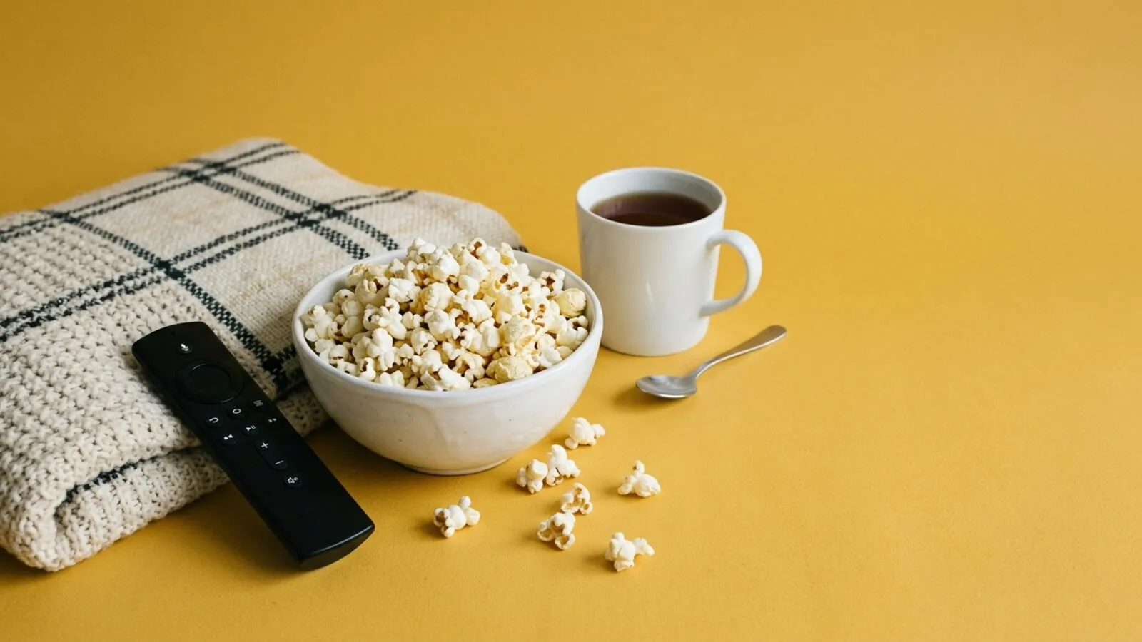 Bol de pop-corn, télécommande noire, plaid écossais crème, tasse de thé et cuillère sur fond jaune