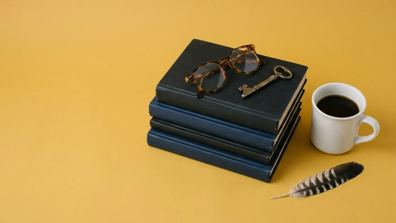 Pile de livres reliés bleu nuit et noirs, lunettes en écaille, vieille clé, tasse de café et plume sur fond jaune