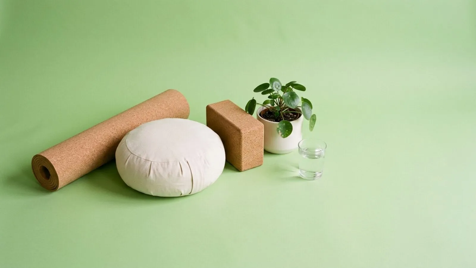 Tapis de yoga en liège roulé, coussin de méditation, brique, plante verte et verre d'eau sur fond vert tendre